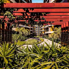 The landscaping at Palm Bellagio is designed to complement the architecture. This view through a red pergola frames the building's unique facade, showing how we integrate green spaces and structural elements to create a cohesive environment.