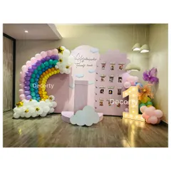 A magical rainbow and cloud-themed backdrop for a first birthday. The setup includes a balloon rainbow, a "My First Year" photo display, and a large light-up number one.
