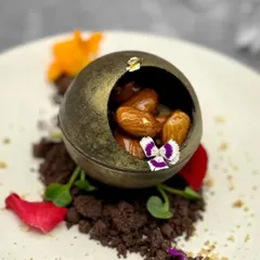 This dessert is a multi-sensory experience. A delicate chocolate sphere holds ginger-spiced almonds, all resting on a bed of chocolate soil and adorned with edible flowers for an artistic presentation.