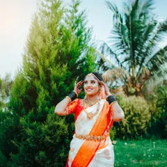 A beautiful outdoor portrait showcasing a traditional South Indian bridal look. The soft, natural sunlight brings out the vibrant orange and white of the saree and the warmth of the gold jewelry, capturing a moment of pure joy.
