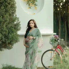A portrait of grace and tradition. The mother-to-be, in a lovely floral saree, holds her baby bump, standing next to a vintage bicycle in a garden setting. This image is a beautiful blend of classic and contemporary elements.