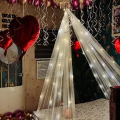 A cozy and romantic room decoration. This setup includes a fairy light canopy over the bed, and the ceiling is filled with metallic pink and red heart-shaped balloons.