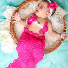 An adorable little mermaid! This baby girl is all smiles in a bright pink, hand-knitted mermaid costume, looking happy and playful in her basket.