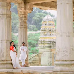 A wide shot of a couple posing amidst the stone pillars of a historic temple, with the gopuram visible in the background. This composition emphasizes the grandeur of the location.
