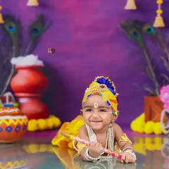 A happy baby dressed as Little Krishna, smiling while holding his flute. The vibrant purple backdrop with peacock feathers and marigolds creates a festive and joyful atmosphere.
