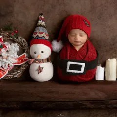 Santa's tiniest helper. A newborn dressed in a little Santa suit, sleeping on a wooden shelf next to a snowman and candles.