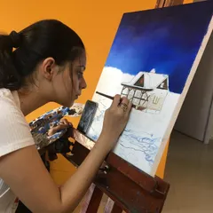 A student carefully works on her acrylic painting of a cozy, snow covered cabin. You can see her focus as she builds the scene, starting with the deep blue sky and working her way to the finer details of the house.