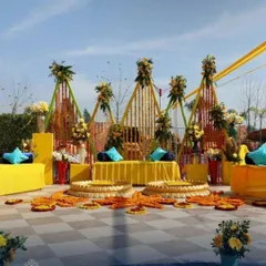 An expansive outdoor Haldi setup with modern triangular frames decorated with marigold strings. The open-air design includes multiple seating areas and traditional 'urli' bowls, perfect for a large gathering.