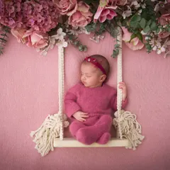 A baby in a cozy pink outfit rests on a macrame swing adorned with an overhead canopy of beautiful pink flowers, a perfect example of our dreamy setups.