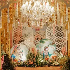 This grand Janmashtami backdrop features intricate white jaali panels, a stunning crystal chandelier, and beautiful illustrations of Krishna. The warm lighting and floral arrangements create a truly heavenly ambiance.