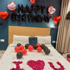 An intimate room decoration for a birthday. The setup includes "Happy Birthday" foil balloons, heart-shaped balloons, and an "I Love You" message written with rose petals on the bed.