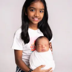 A proud big sister holds her swaddled newborn sibling in this classic studio portrait. Her bright smile shows her excitement at her new role.