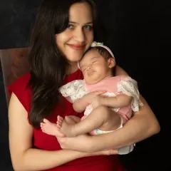 A classic portrait of a mother in a red dress, smiling warmly as she holds her sleeping newborn angel against a simple dark background.