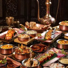 A lavish spread of Awadhi cuisine, featuring biryani, kebabs, and curries in copper and brass vessels. The dark, opulent styling with an antique ewer evokes the royal heritage of Lucknowi food.