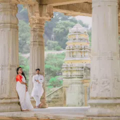 A wide shot of a couple posing amidst the stone pillars of a historic temple, with the gopuram visible in the background. This composition emphasizes the grandeur of the location.