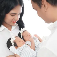 A close-up shot showing the parents' loving expressions as they hold their sleeping twin daughters. The bright, high-key lighting creates a pure and angelic feel.