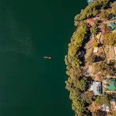 A top-down aerial shot of the shoreline of Naukuchiatal, contrasting the deep green water with the lush forest and houses. A lone boat adds a sense of scale and tranquility to this beautiful Uttarakhand landscape.