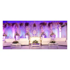 An exceptionally detailed and luxurious reception stage. The intricate white and gold carved backdrop, illuminated by soft purple lighting and adorned with flowers, provides a truly regal setting for the couple.