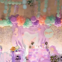A full stage setup for a mermaid party, featuring a scalloped dessert bar, character cutouts, and a ceiling of hanging balloons that look like bubbles and seaweed.