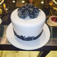 A striking and minimalist cake design. The clean white fondant is contrasted with a cluster of black-dusted roses on top and a border of chocolate sprinkles, making for a modern and sophisticated statement.
