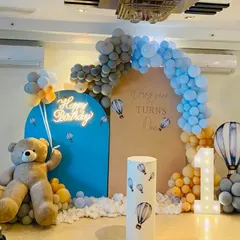 A whimsical 'hot air balloon and teddy bear' theme for a first birthday party. This setup includes a custom arched backdrop, a light-up number '1', and a life-sized teddy bear, all tied together with a soft blue, grey, and yellow color palette for a picture-perfect celebration.