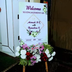 A classic welcome sign for an engagement ceremony, clearly stating the couple's names and event date. The simple floral arrangement at the base adds a touch of freshness and celebration.