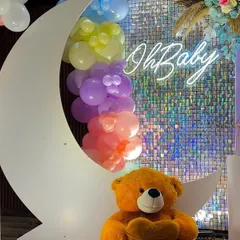 A close-up of the crescent moon prop, filled with pastel rainbow balloons and set against a silver shimmer wall. The "Oh Baby" neon sign and a cute teddy bear complete this perfect photo opportunity.