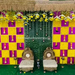 A vibrant setup for an Upanayanam or sacred thread ceremony. The backdrop is a checkerboard of yellow marigolds and pink fabric, with hanging bells and two traditional chairs for the ceremony.