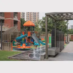 A small water feature and shaded seating are placed adjacent to the play area, allowing for comfortable supervision and sensory play.