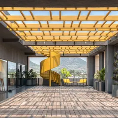 A bold, yellow spiral staircase serves as a sculptural centerpiece on the terrace of the Laurus Labs regional office in Vizag. This open-air space was designed to maximize natural light and offer stunning views of the surrounding hills, creating an inspiring breakout area that encourages movement and connection with the outdoors.