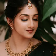A close-up bridal portrait focusing on the bride's flawless makeup and serene expression. The soft lighting beautifully highlights her features and traditional gold jewelry.