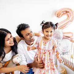 A happy family moment celebrating a second birthday at home. The balloons in the background add a festive touch, but the real story is the shared laughter between the parents and their daughter.