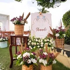 A rustic and charming welcome setup for a "Shaadi," featuring a custom sign, fresh flowers in wicker baskets, and pampas grass accents.