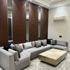 This L-shaped sectional sofa in a soft grey fabric provides ample seating, paired with a striking brass-finished center table. The back wall features a modern design of wood veneer and mirror panels, adding depth to the room.