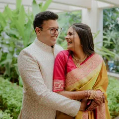 A joyful photo of a couple sharing a laugh during their engagement shoot. Her vibrant yellow and pink saree and his elegant sherwani look stunning against the green foliage.