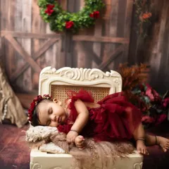 A sleeping baby girl in a rich red dress, resting on an elegant chaise against a rustic barn door backdrop. The Christmas wreath adds a festive touch.