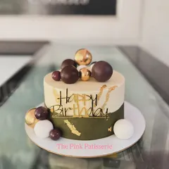 A contemporary birthday cake featuring a unique color block design in olive green and ivory. The cake is adorned with chocolate and gold spheres, gold leaf accents, and a custom acrylic topper.