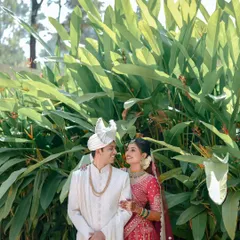 A classic portrait of a Maharashtrian couple sharing a look of love, framed by lush green leaves. The bride's red lehenga and the groom's white sherwani create a beautiful contrast.