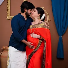 A loving portrait of a couple in a traditional setting. The mom-to-be is radiant in a red and purple saree, while her partner complements her in a dhoti. This setup is designed to celebrate your journey with cultural richness and elegance.
