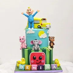 A two-tier Cocomelon cake for Demira's birthday, featuring JJ and the yellow school bus on top of a vibrant, character-filled design.