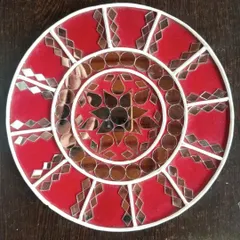 This sample piece shows a more geometric approach to Lippan art. The bold red background contrasts with the white clay work and reflective mirrors, demonstrating a different style you can explore in the workshop.