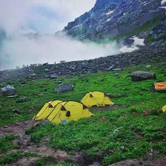 Tents pitched on a vibrant green meadow, with misty clouds rolling through the mountain pass. This is a scene from a group trek, where the journey is as beautiful as the destination.