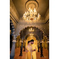 A couple shares a dance in a grand hall with beautiful arches and chandeliers. Their matching yellow outfits complement the golden lighting perfectly.