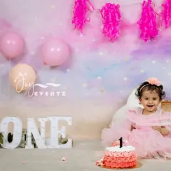 A beautiful pastel-themed cake smash photoshoot for a baby girl's first birthday. The setup includes a light-up "ONE" sign, pink balloons, and a lovely ombre cake, creating a dreamy and celebratory atmosphere.