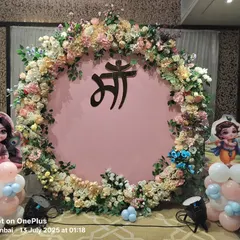 A beautiful baby shower backdrop with a large floral ring surrounding the 'Maa' (Mother) symbol in Hindi. The setup is flanked by cute Krishna and Radha cutouts on balloon pillars.