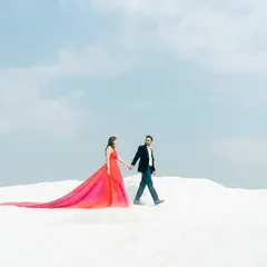A wide-angle shot from a pre-wedding photoshoot, capturing the couple walking hand-in-hand against a vast, minimalist landscape. The long trail of the gown adds a sense of scale and drama to this romantic portrait.