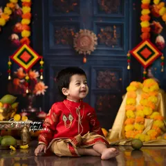 The joy of celebration shines in this little boy's smile. Dressed in a festive red kurta and dhoti, he is surrounded by a traditional setup of marigold garlands and brass lamps.