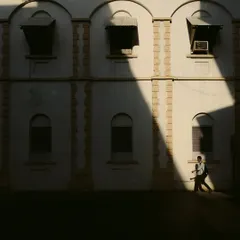 A play of light and shadow on a building's facade, with a lone figure walking into a sliver of sunlight. This minimalist composition uses strong diagonal lines and contrast to create a dramatic and artistic street photograph.