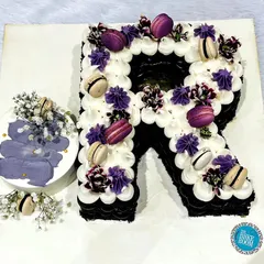 A personalized monogram cake. This "R" shaped cake is decorated with purple and white buttercream swirls and macarons, and comes with a matching mini bento cake.