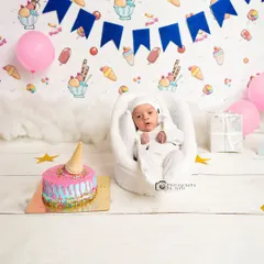 An adorable ice cream-themed milestone shoot. This newborn is surrounded by sweet decorations, including a colorful fake cake and an ice cream-patterned backdrop.